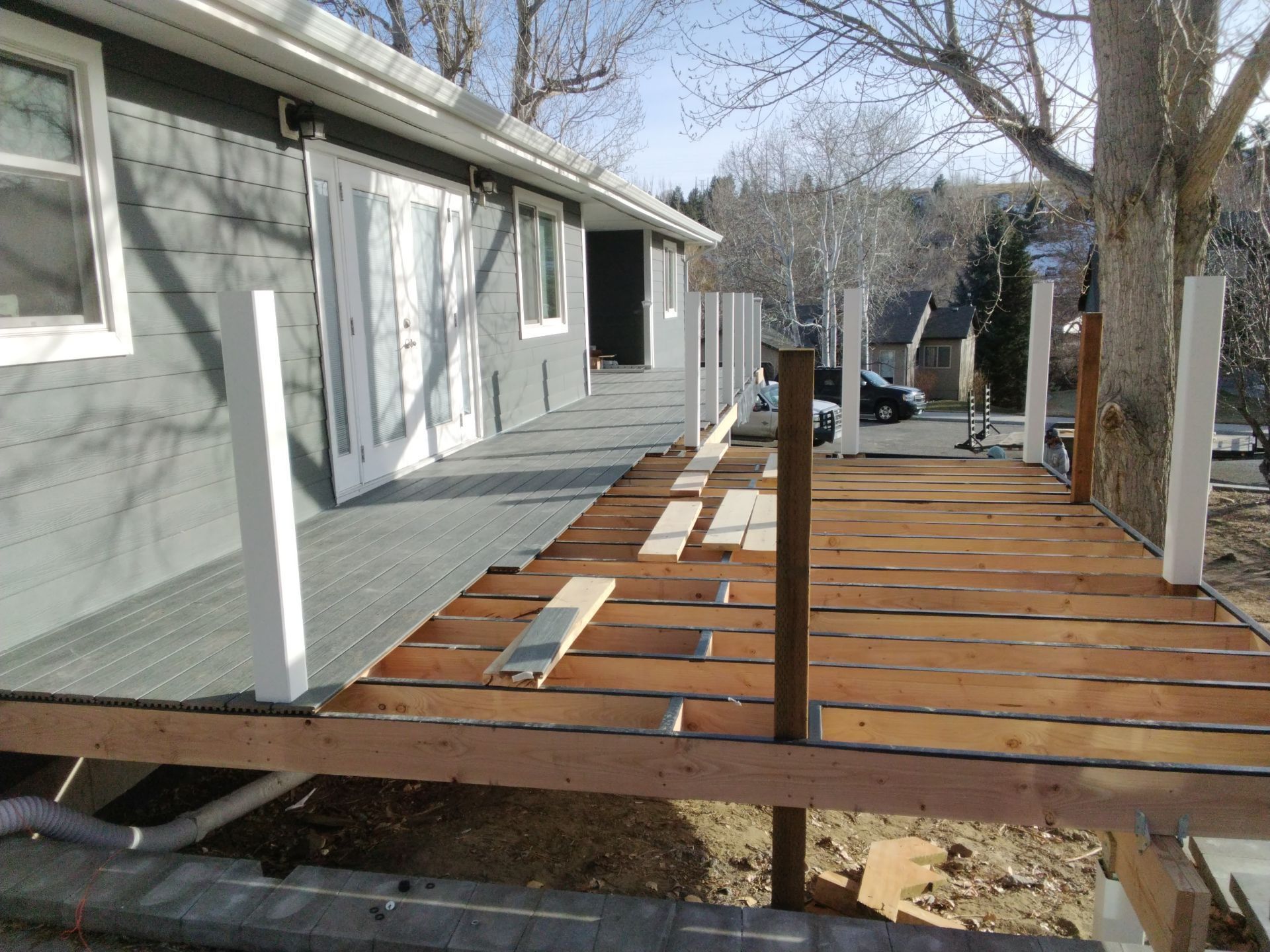 Deck construction in progress; wooden deck boards being installed with white railing posts, alongside a gray house.