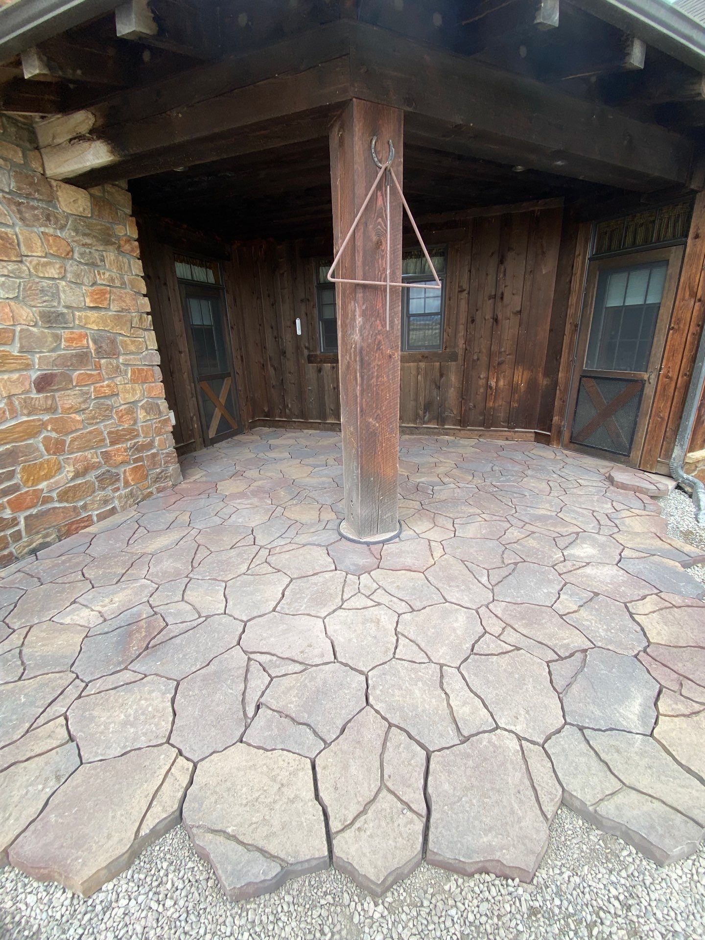 Stone patio under wooden porch with central support beam. Brown stone and wood.