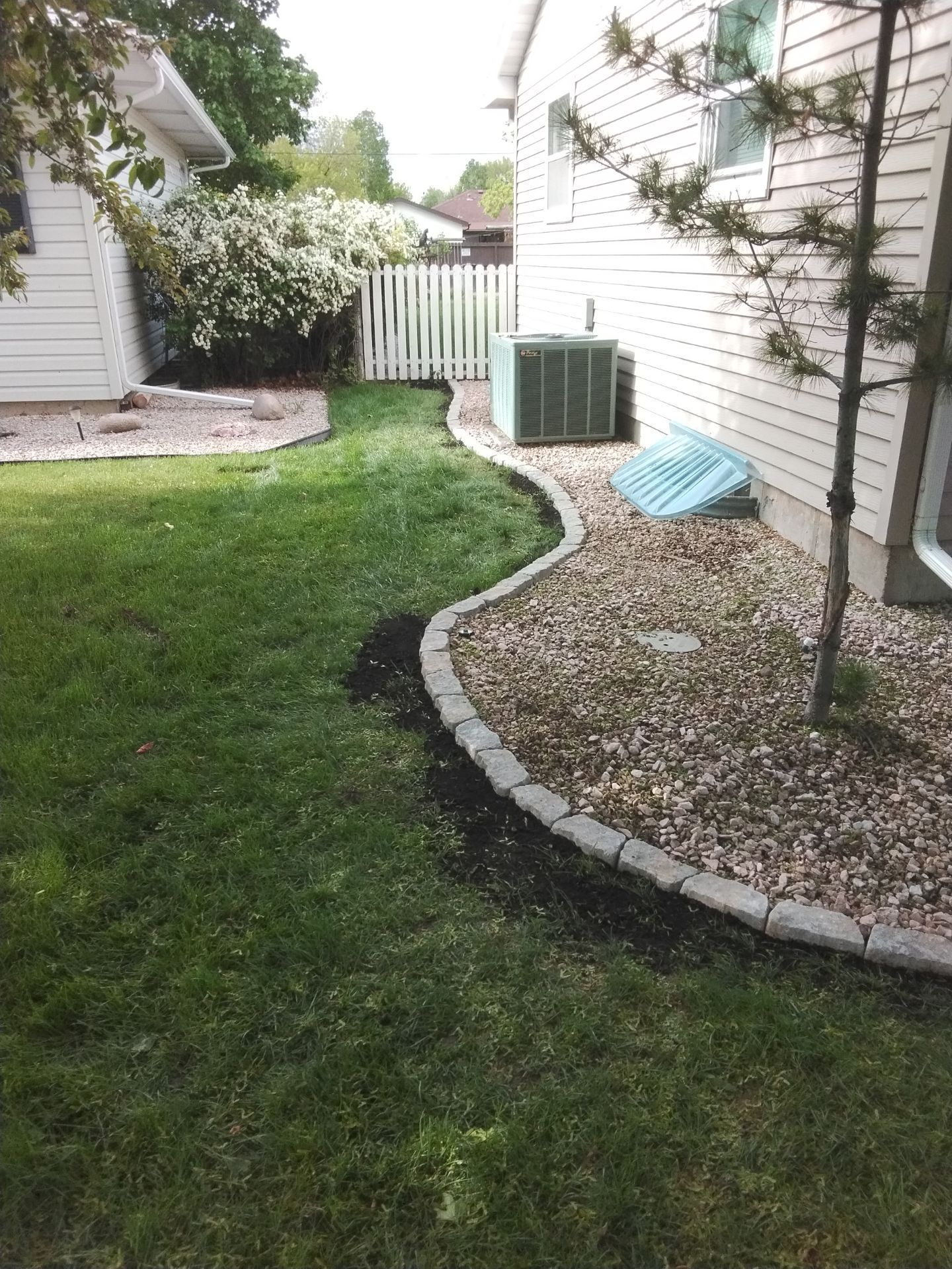 Lawn edged with gray pavers curves along the side of a house, separating grass and gravel landscaping.