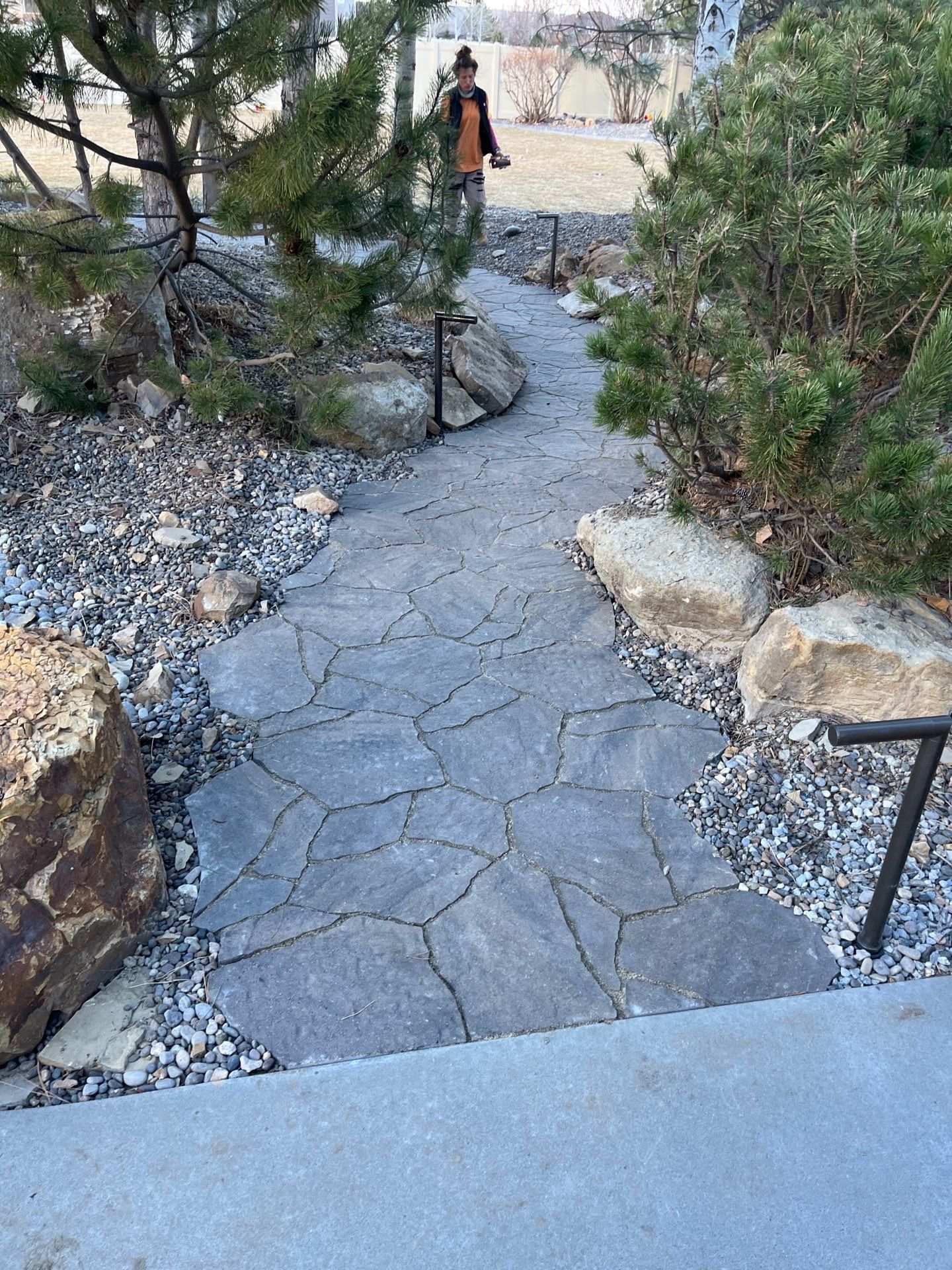 A stone pathway winds through a garden with large rocks and greenery, person in the background.