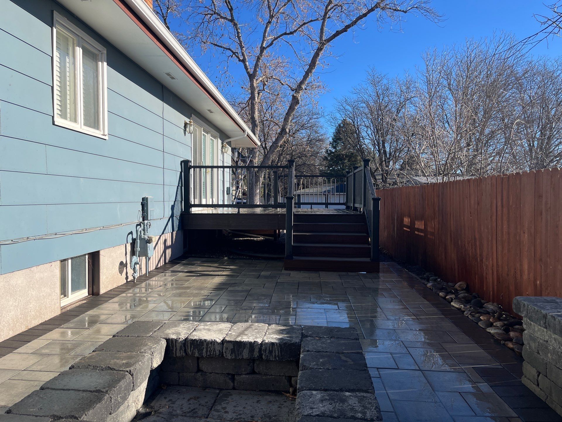 Blue house with a paved patio, dark deck, and wooden fence on a sunny day.