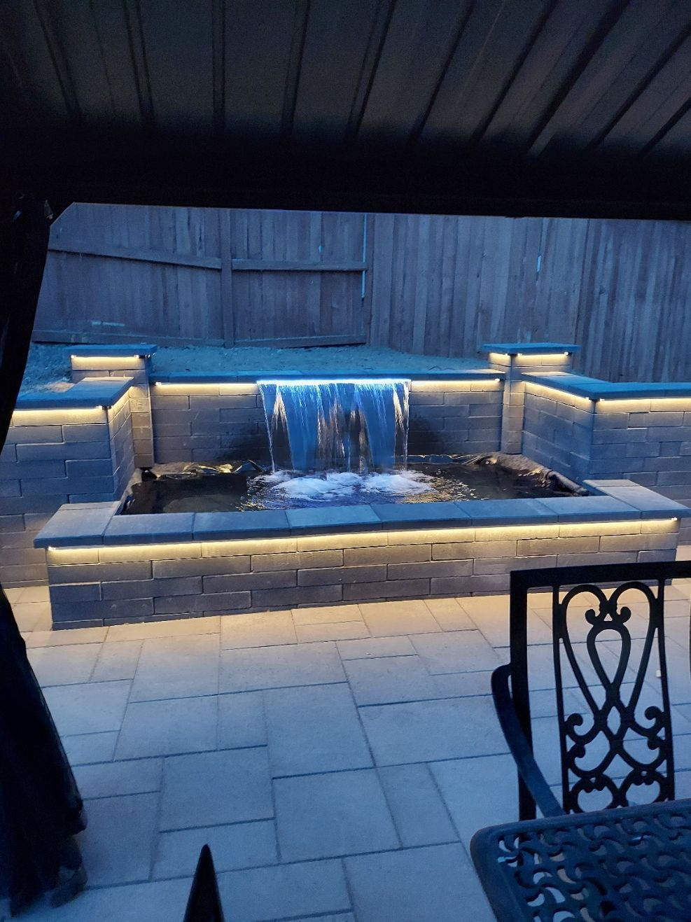 Water feature with waterfall cascading into a pond, lit by cool-toned lights, on a patio.