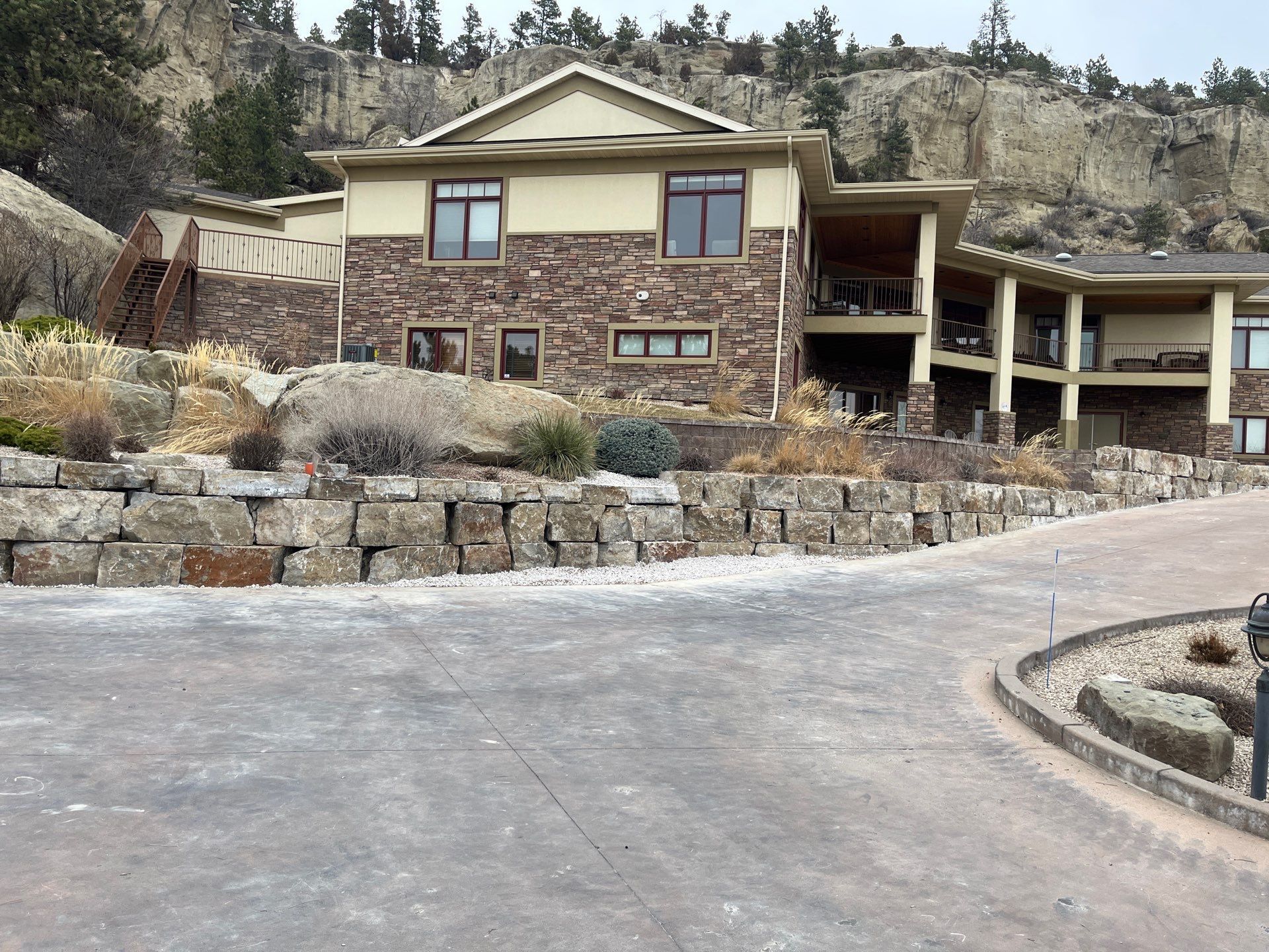 House with stone facade and retaining wall, set against a rocky hillside, visible driveway.