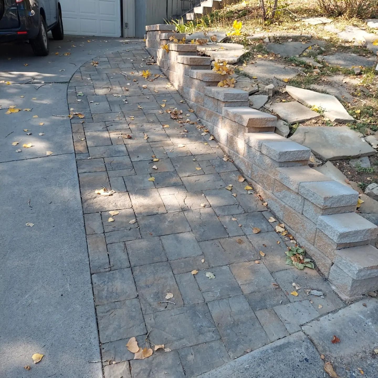 Brick pathway next to retaining wall and driveway. Scattered leaves, sunlight.