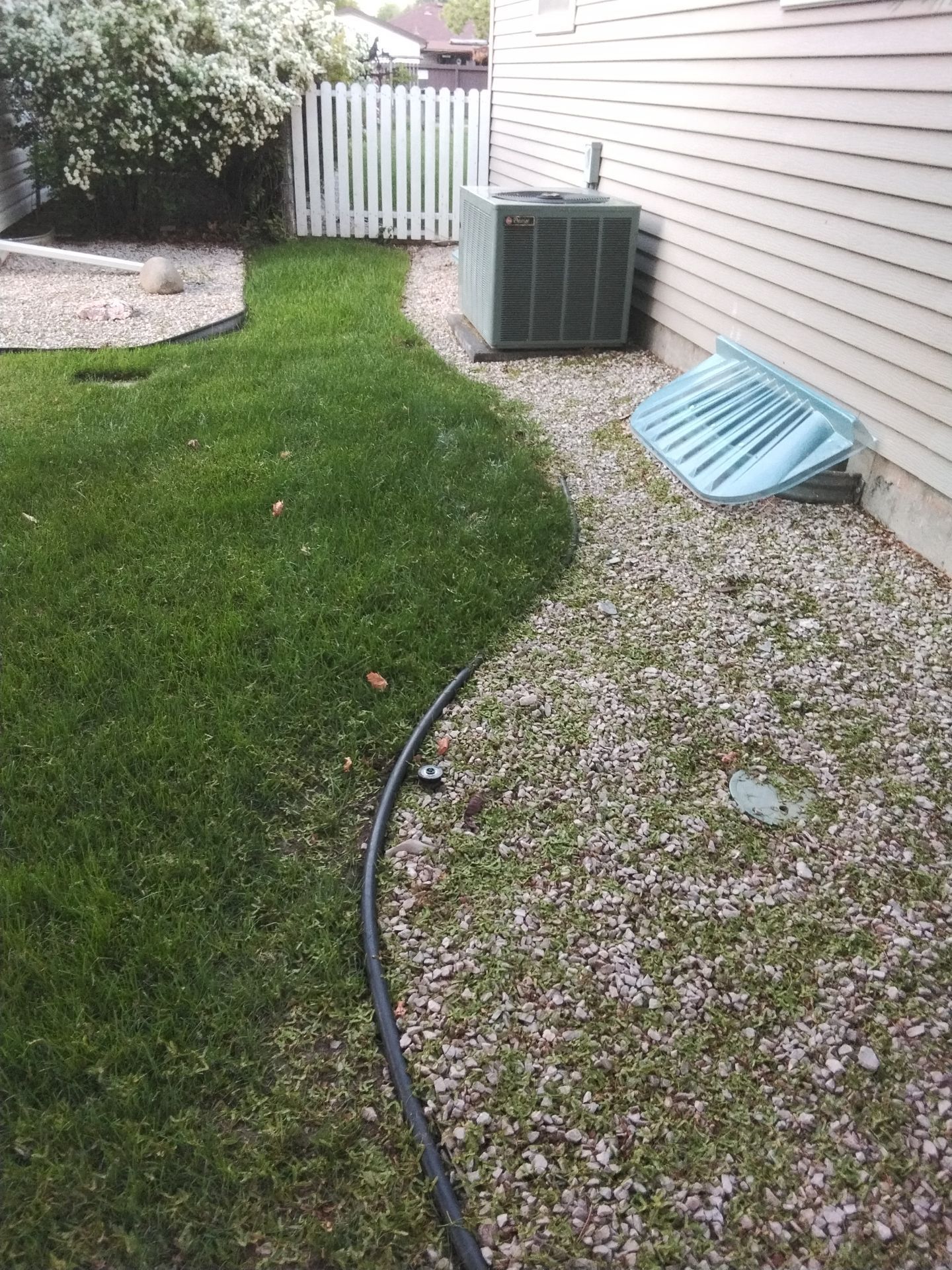 Green grass meets gravel pathway beside a house and air conditioner. White fence in background.