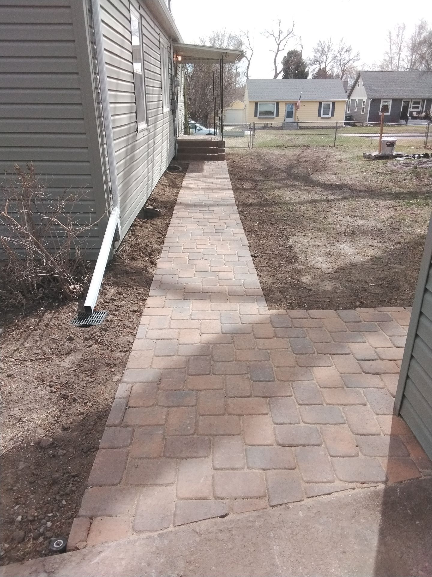 Brick walkway next to a light gray house, leading to a porch. Bare yard on the right.