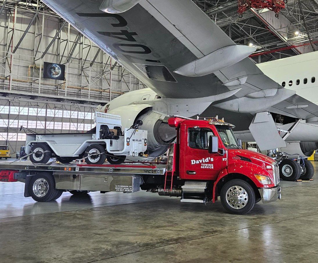 A small vintage car loaded on a red tow truck inside a large hangar with a plane overhead.