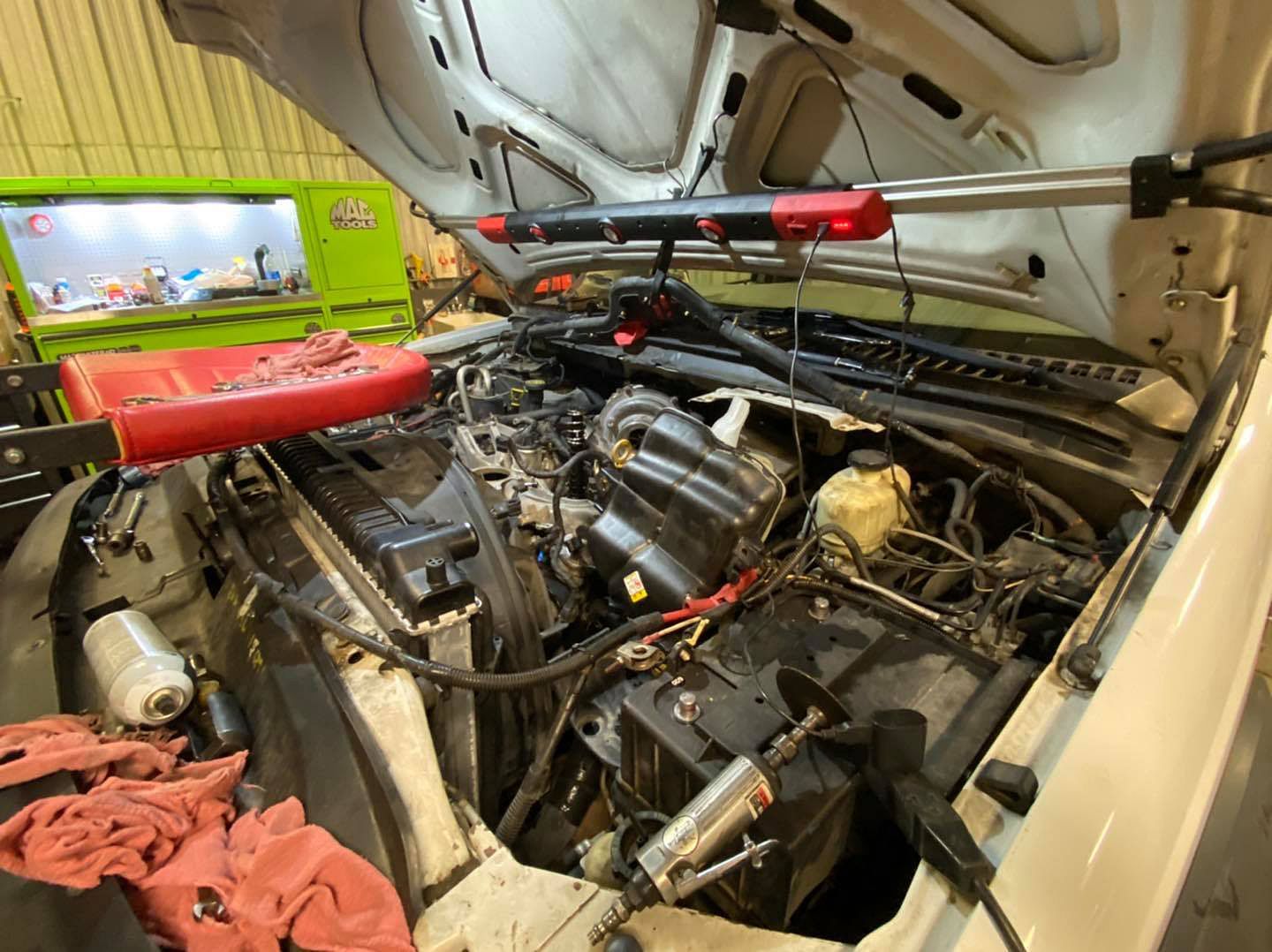 Car engine bay with hood open, tools and lights, likely in a garage.