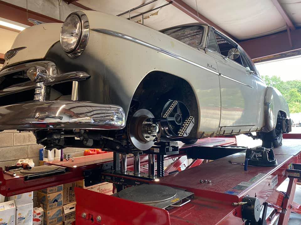 Classic car on a lift for repairs. White and silver, with exposed front wheel. In a garage.