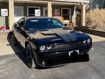 A black Dodge Challenger with a racing stripe parked on a driveway in front of a building with porch seating.