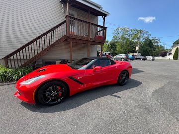 A bright red Chevrolet Corvette convertible parked on a gravel lot next to a building with an exterior wooden staircase.