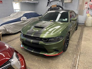 An olive green Dodge Charger with black racing stripes parked in a garage, with another car covered in a gray tarp.