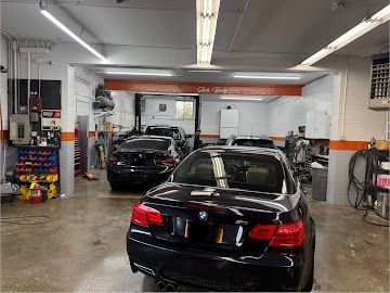 An auto repair shop interior showing two black cars parked in bays with concrete floors and bright overhead lighting.