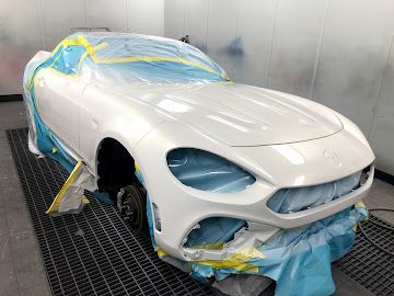 A white sports car covered in protective tape and plastic sheeting inside an auto body shop spray booth.