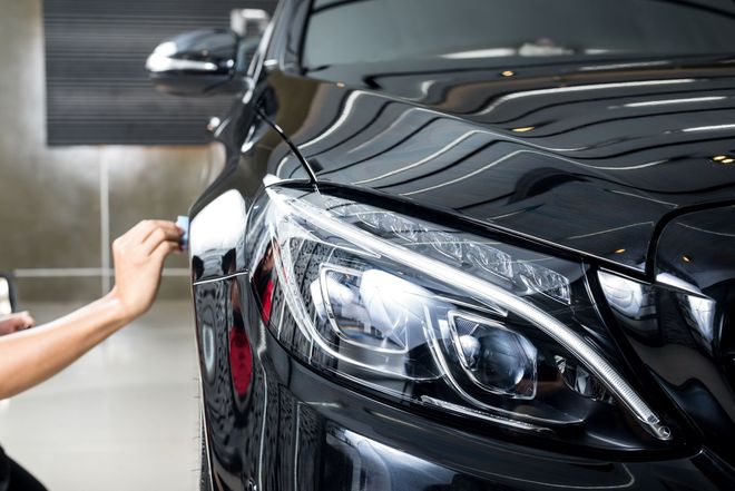 A person's hand applying a protective film to the headlight of a glossy black car in a garage setting.