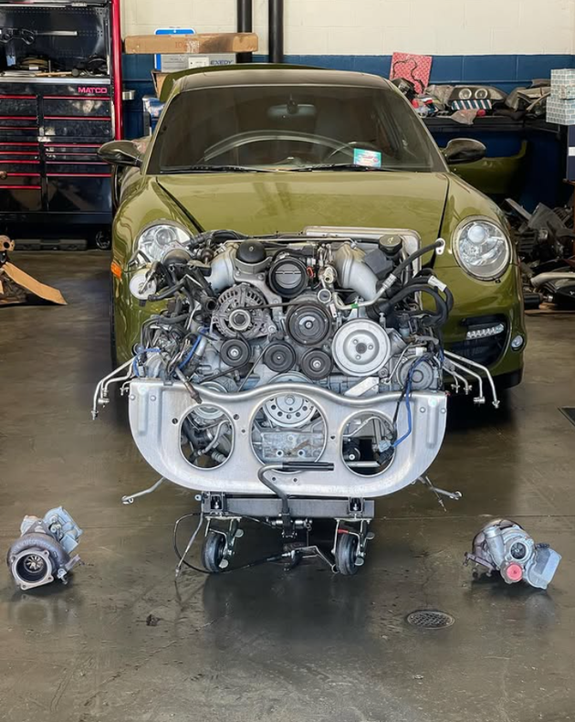 Green car in a garage with its front bumper removed, exposing the engine and parts on the floor