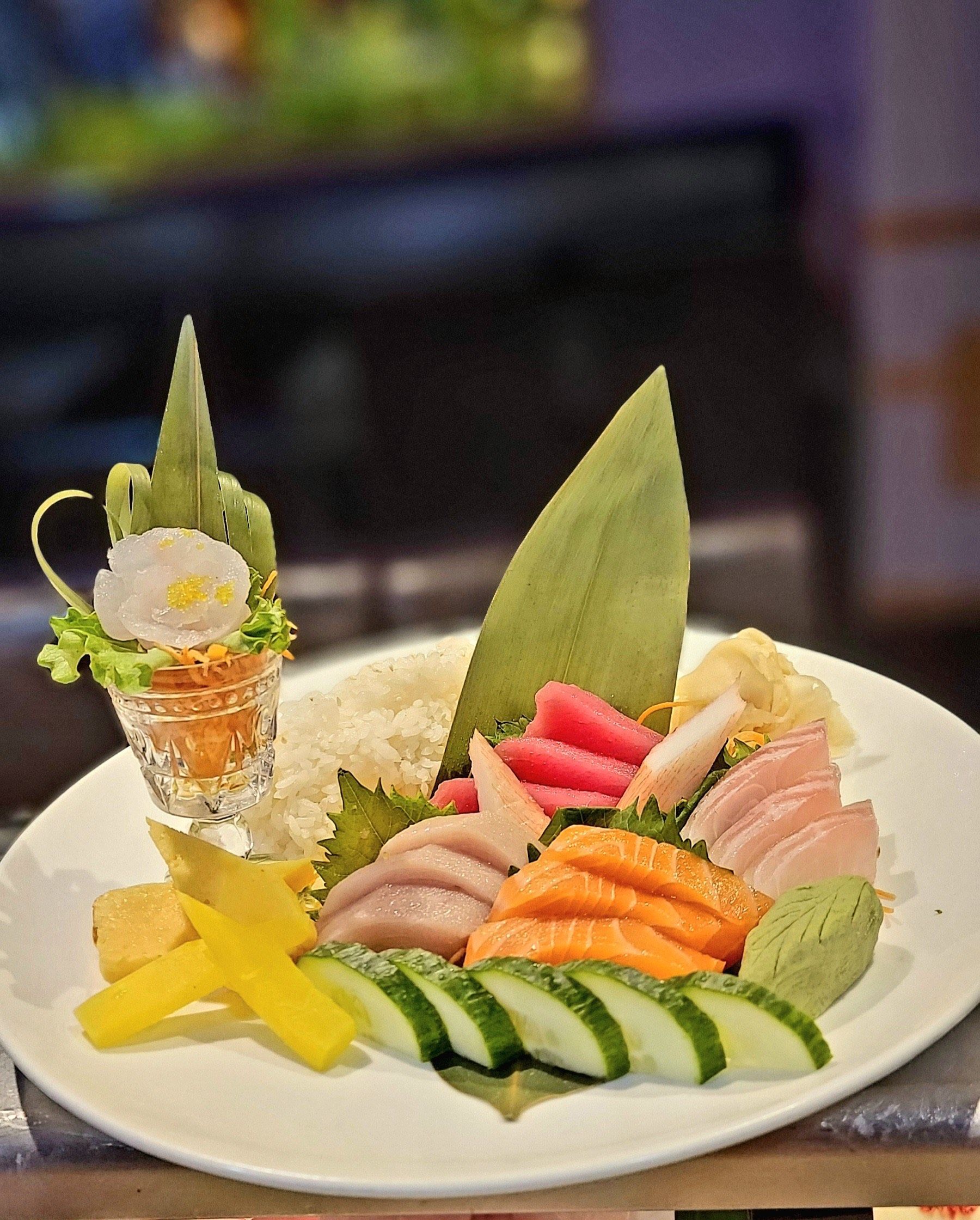Sushi platter with various sashimi, cucumber slices, wasabi, and a decorative floral arrangement.