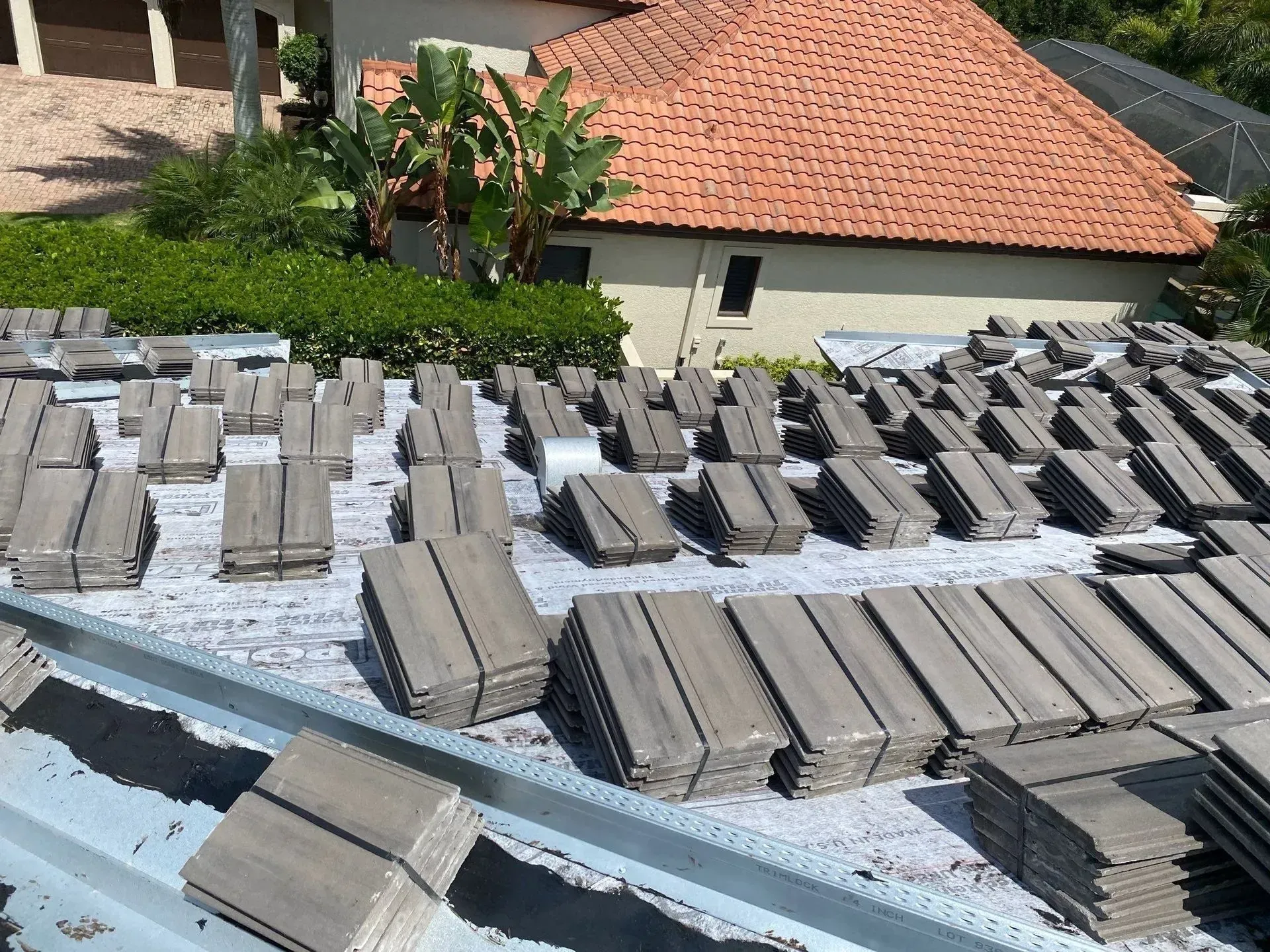 Stacks of gray roof tiles on a rooftop with a red-tiled house and greenery in the background