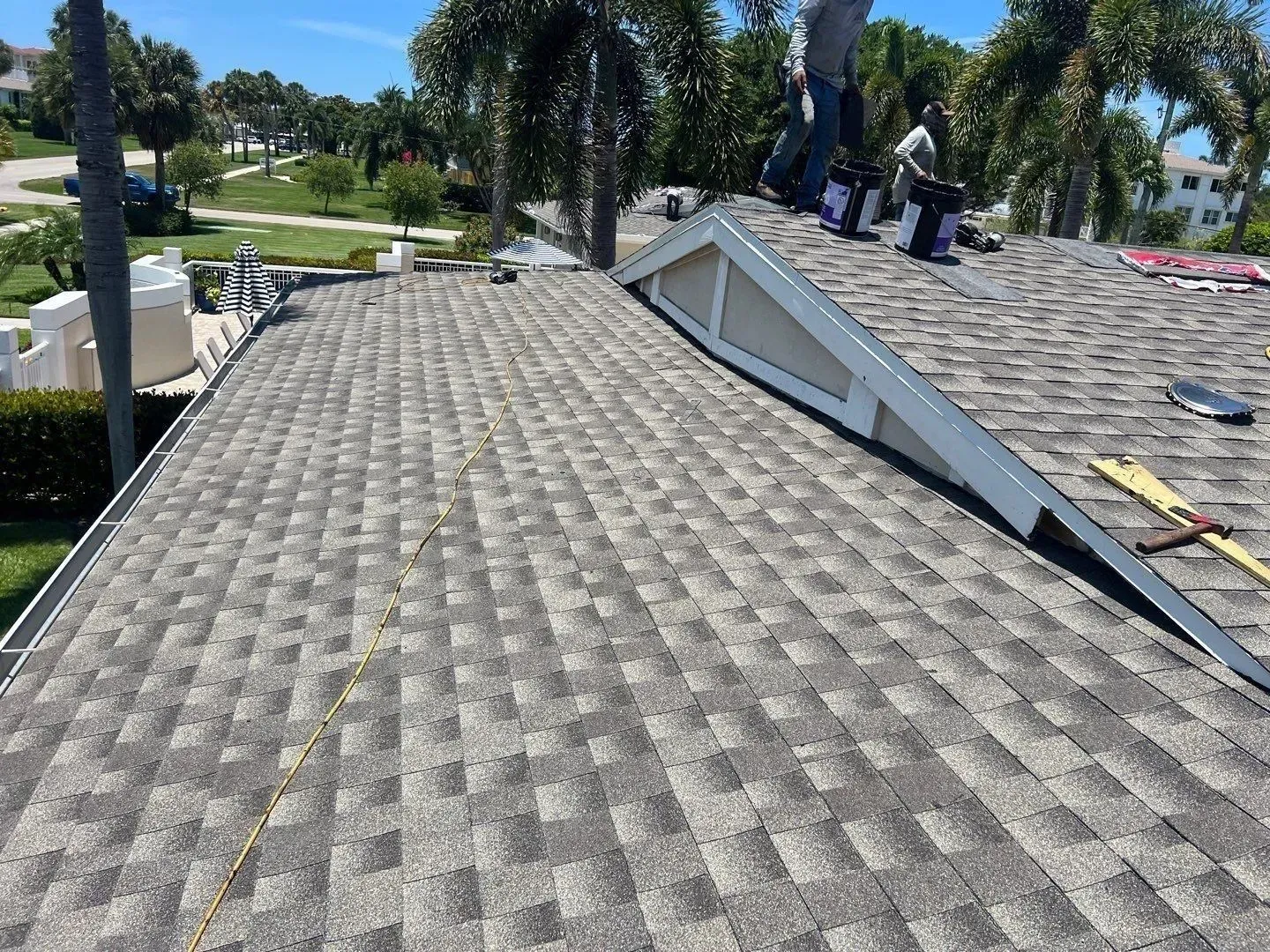 Roof with gray shingles and a white dormer, with people working in the background under palm trees.