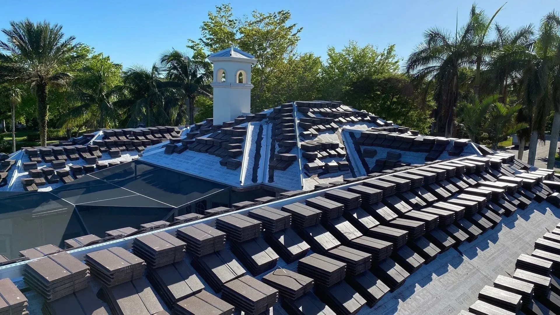 Blue-tiled roof with rows of dark roof windows around a small white tower, surrounded by palm trees.