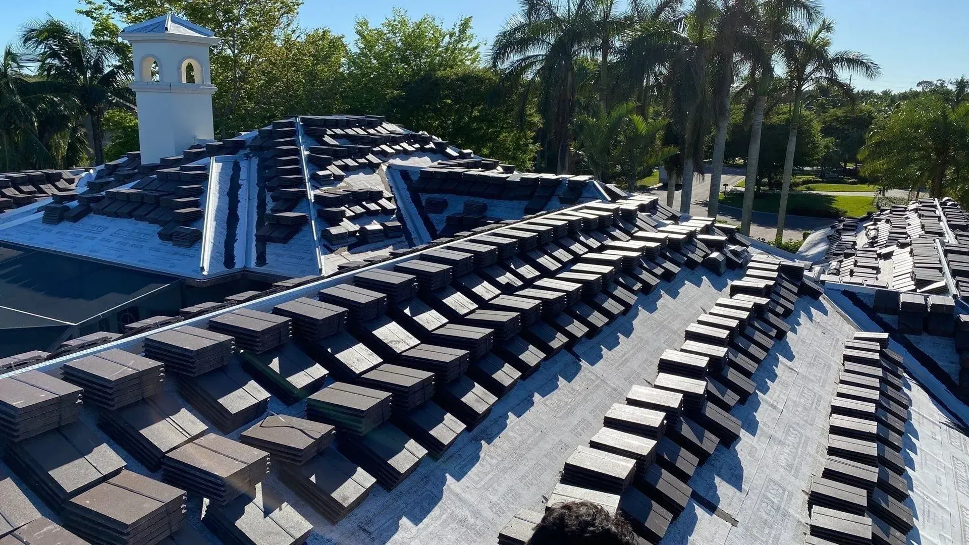 Rows of dark roof tiles laid out on a sunlit residential roof beside palm trees and a white tower.