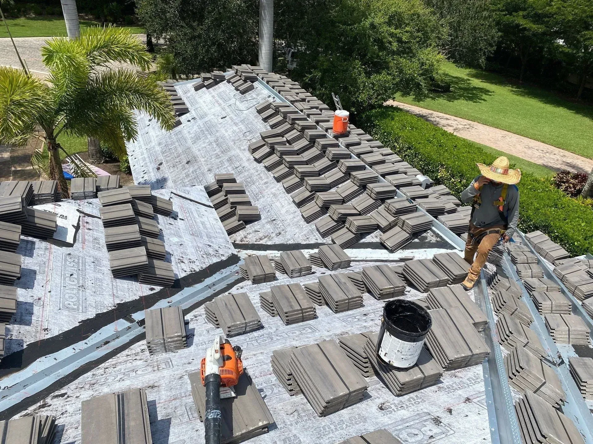 Workers installing gray roof tiles on a house roof, with one person wearing a straw hat nearby.