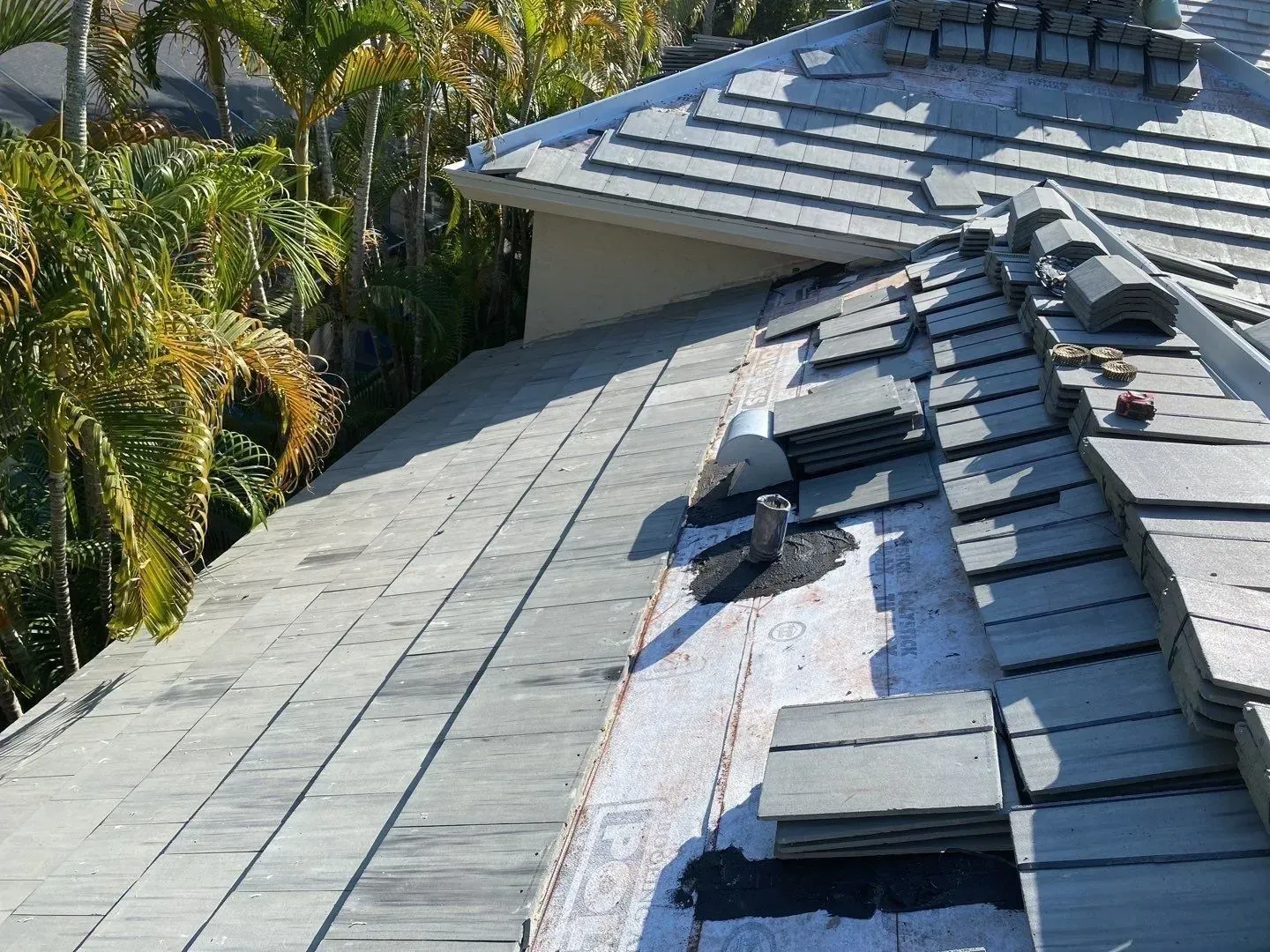 Roof under repair with dark shingles and exposed underlayment beside a palm tree