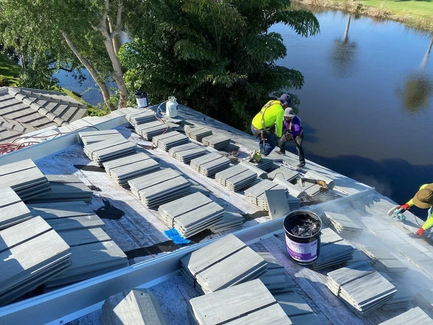 Workers painting a roof beside a river, with rows of gray panels and a bucket of paint