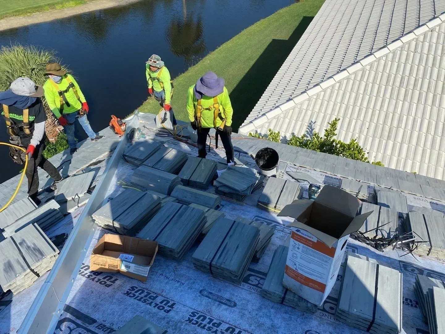 Workers in neon safety gear installing roofing tiles on a sloped roof beside water