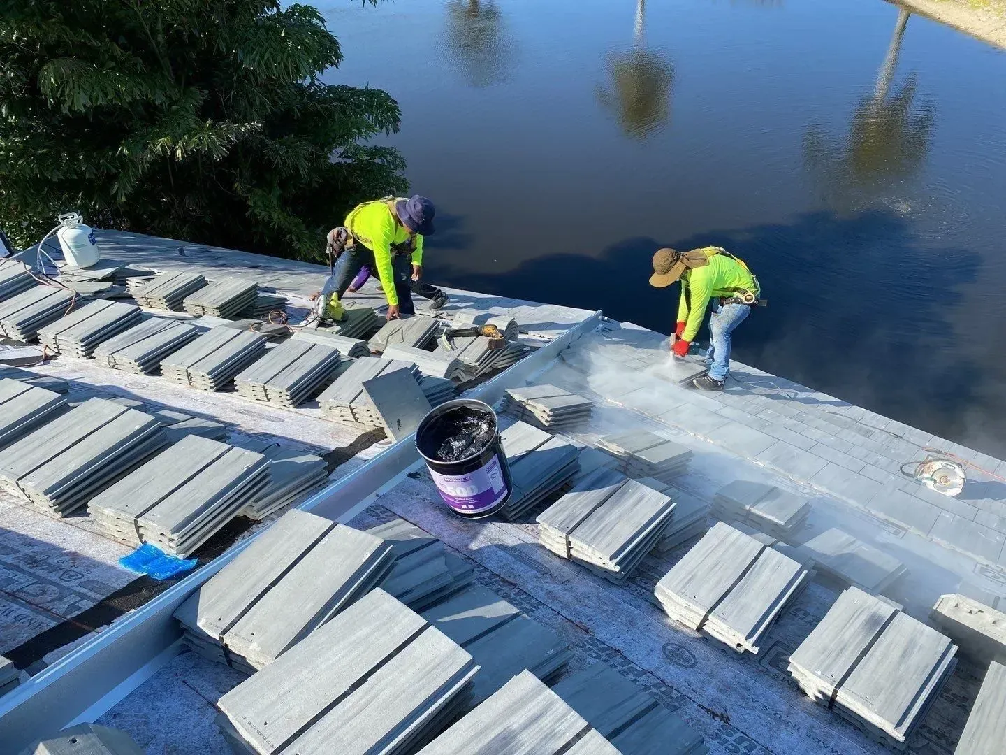 Workers in neon safety gear spread white tiles on a rooftop by water