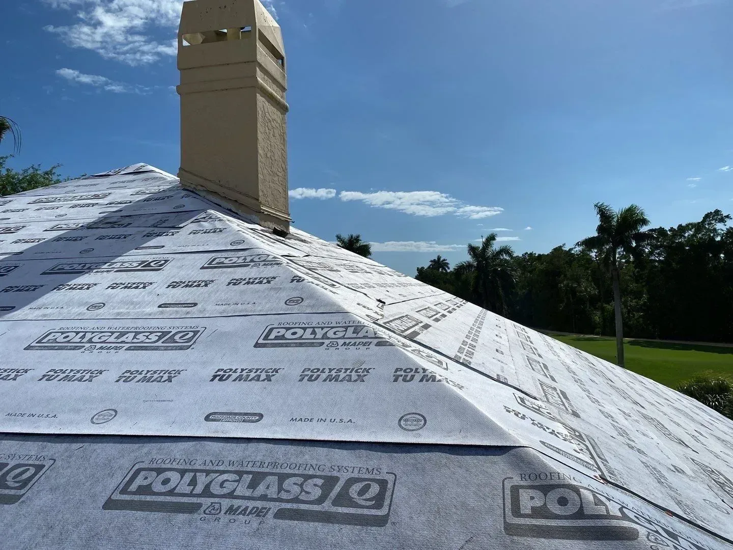 Roof under construction with white underlayment, tan chimney, and palm trees under a blue sky