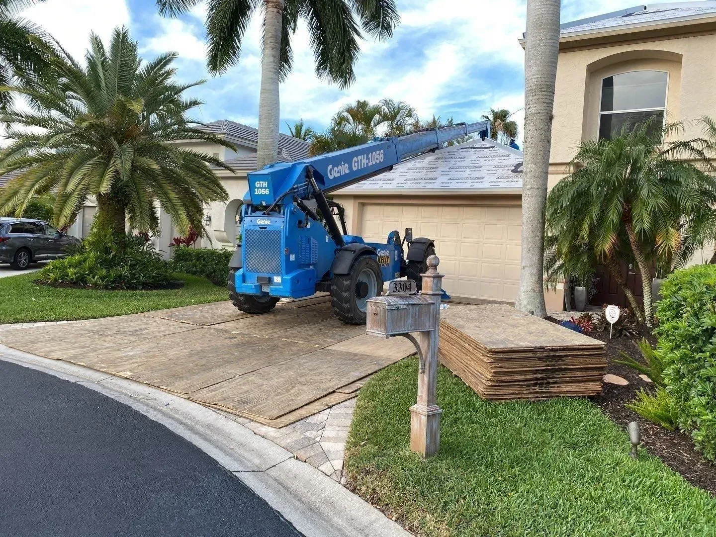 Blue boom lift parked in a driveway beside a house, with plywood stacked near the garage.