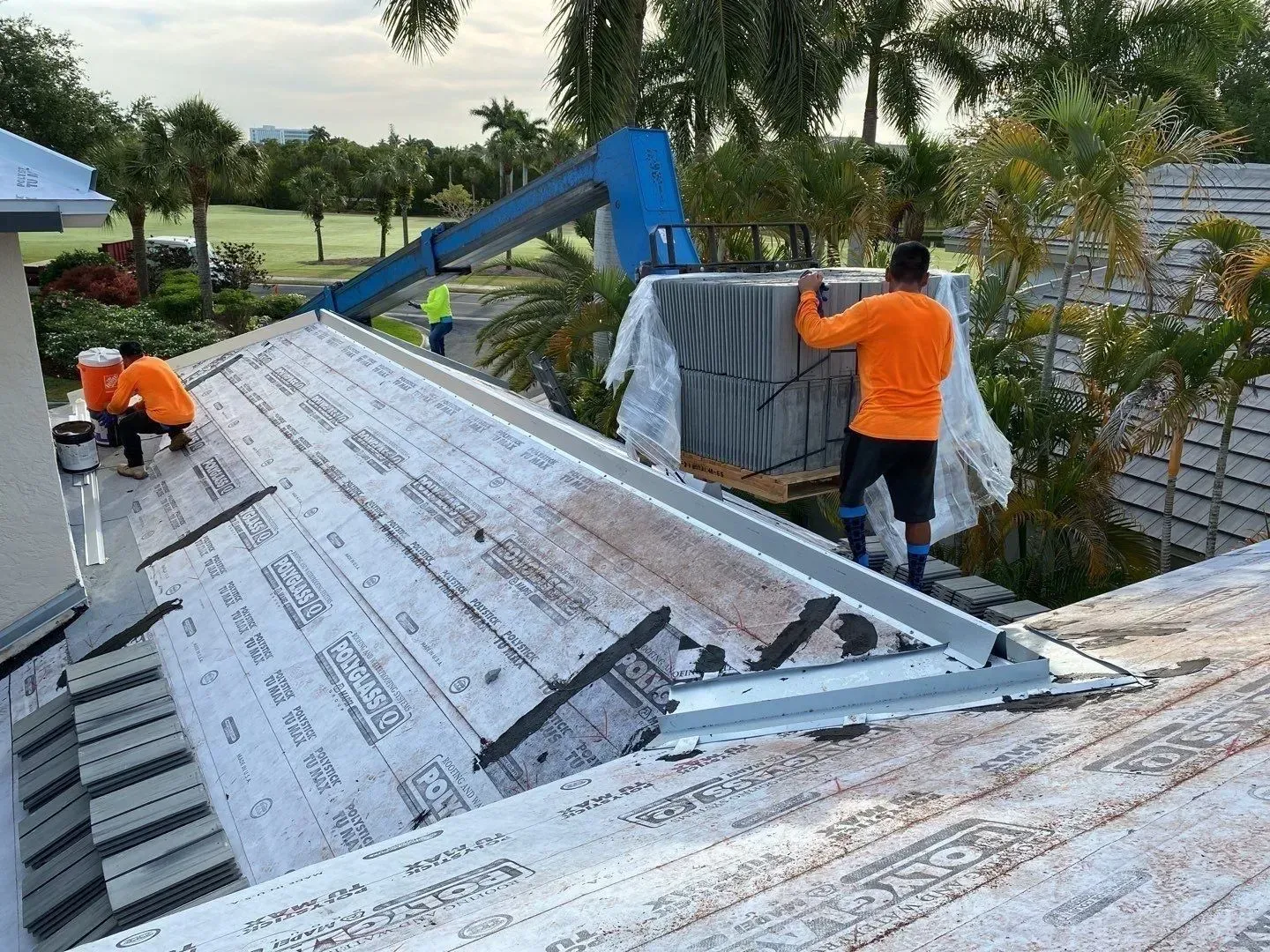 Workers in orange safety shirts installing roof panels with a crane on a sloped roof.