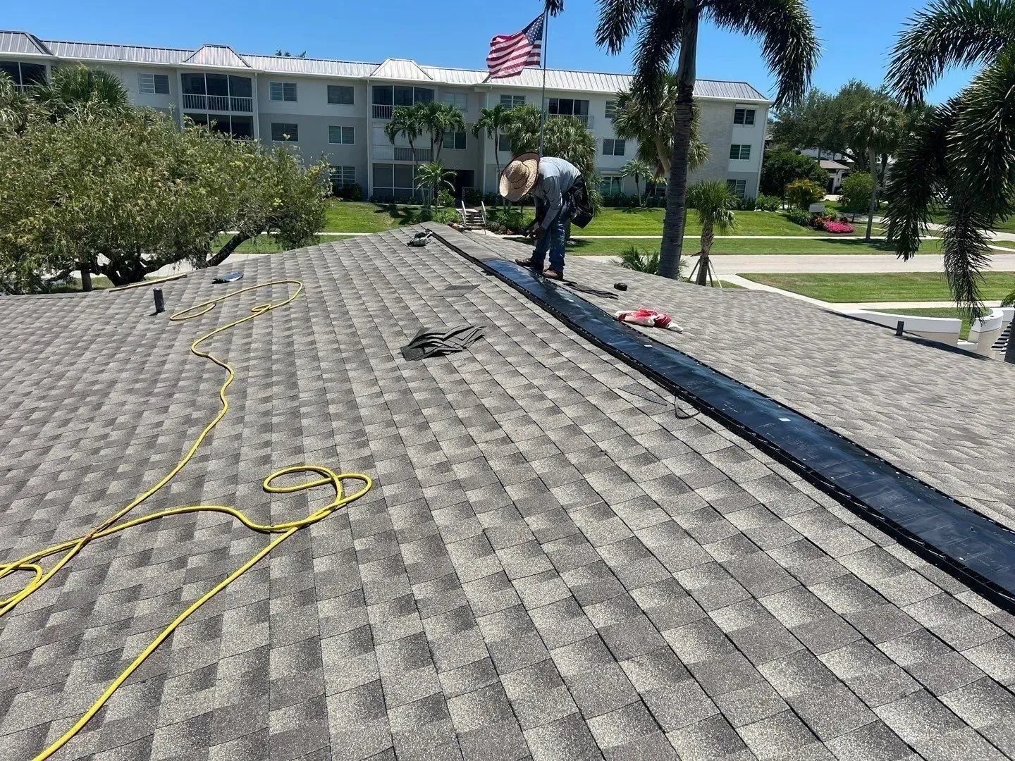 Workers on a roof repairing shingles beside a park and an American flag