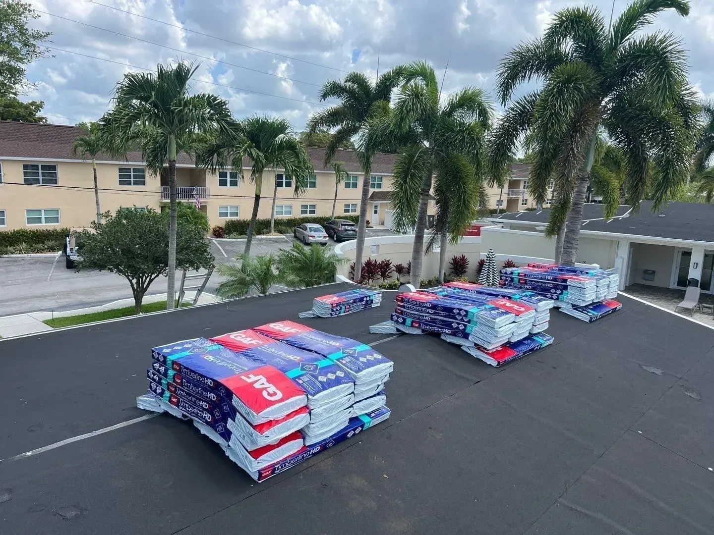 Pallets of red, blue, and white roofing shingles stacked in a parking lot with palm trees and buildings nearby