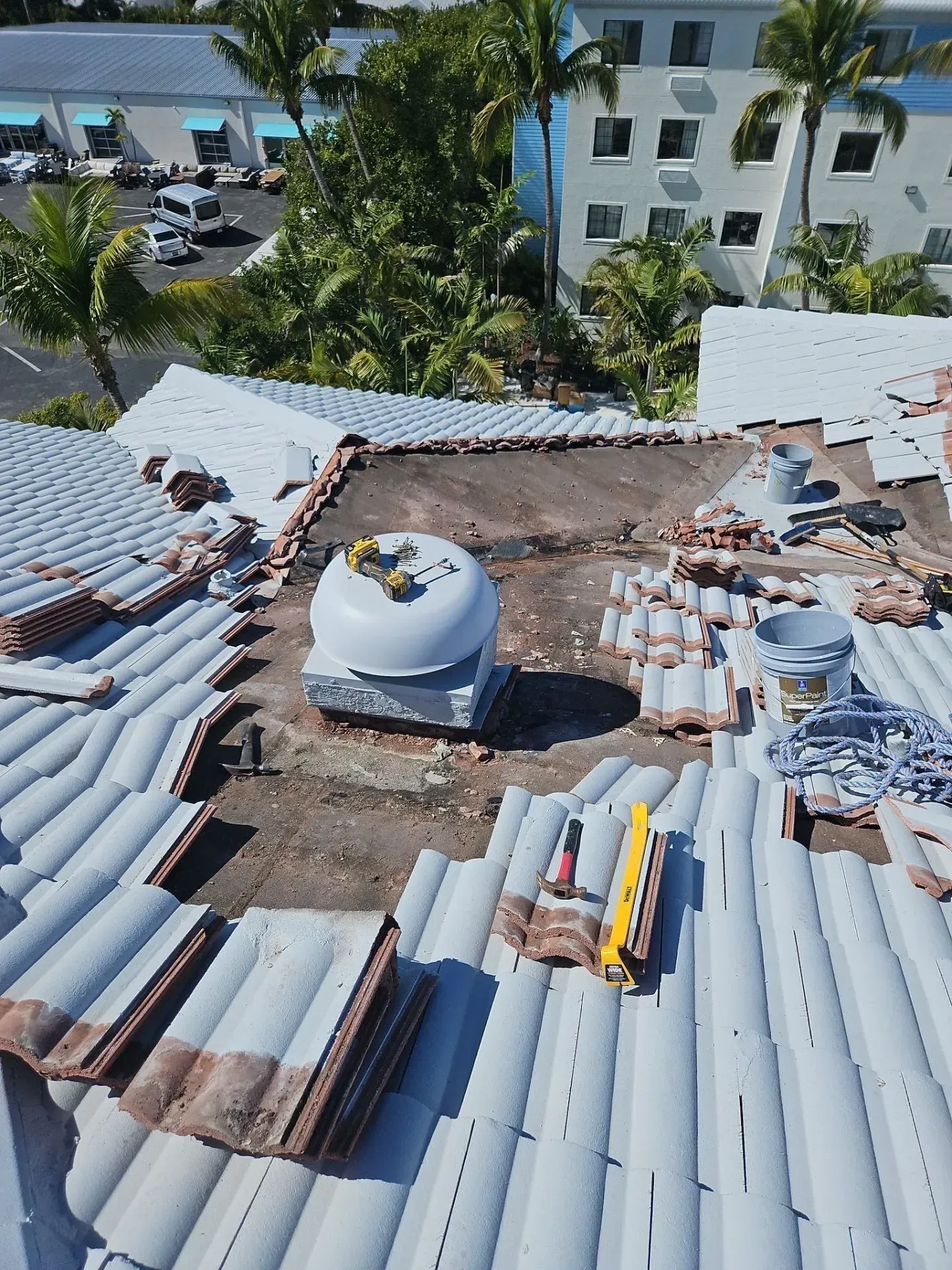 Rooftop under construction with stacks of roofing tiles and a white dome vent, overlooking palm trees and buildings