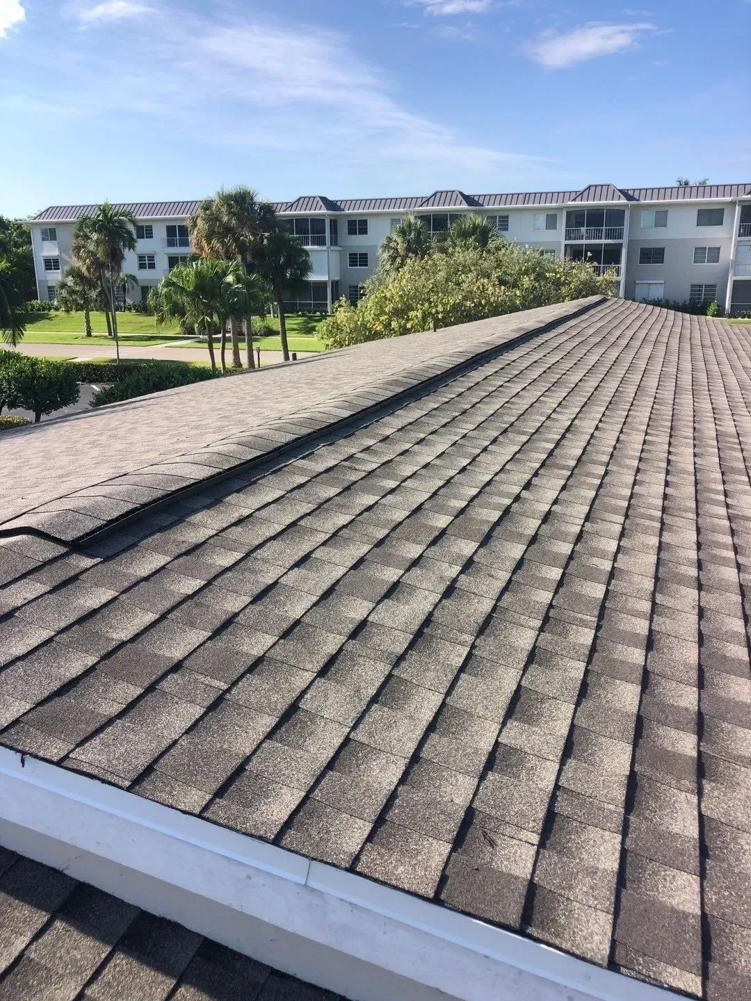 Rooftop with brown shingles overlooking white apartment buildings, green lawn, and blue sky