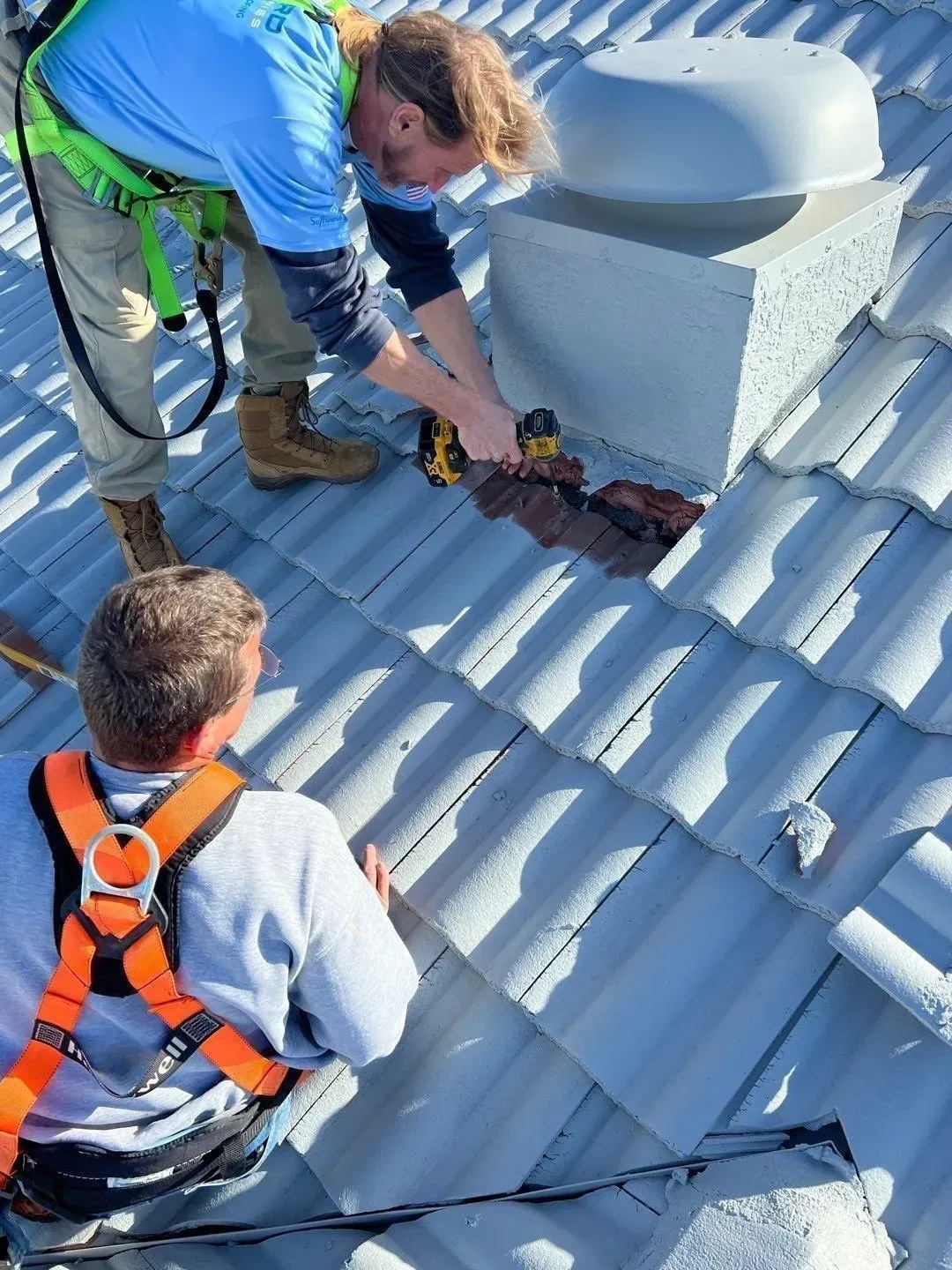 Two workers repairing roof shingles around a vent stack on a steep metal roof