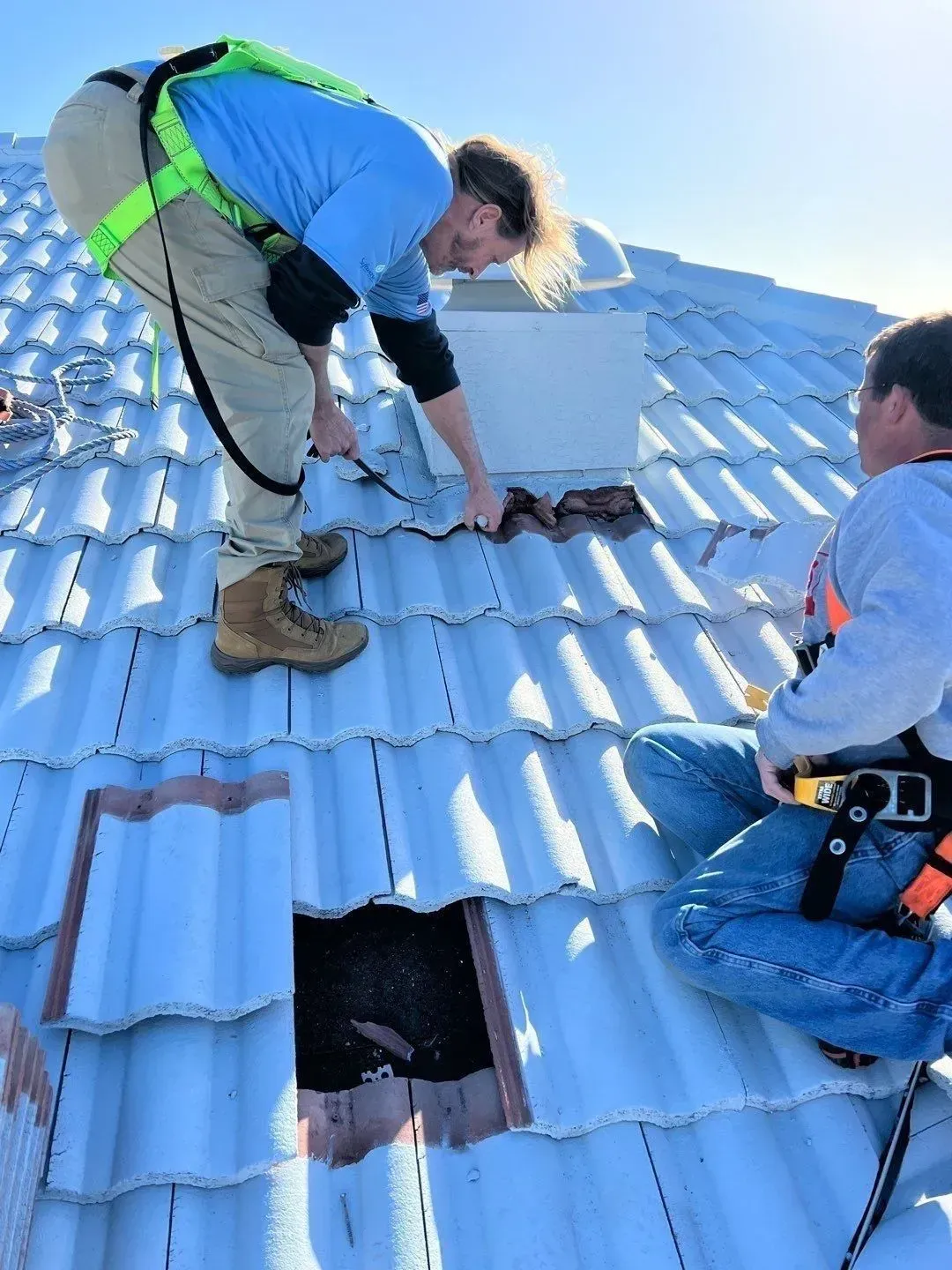 Two workers repairing a blue tiled roof, one kneeling over a damaged section on a sunny day