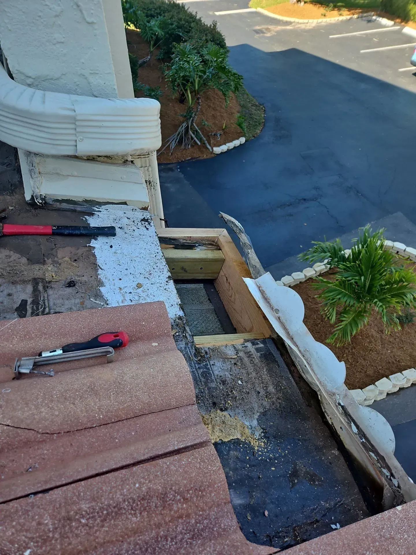 Roofing repair with tools on shingles beside a damaged edge overlooking a parking lot below