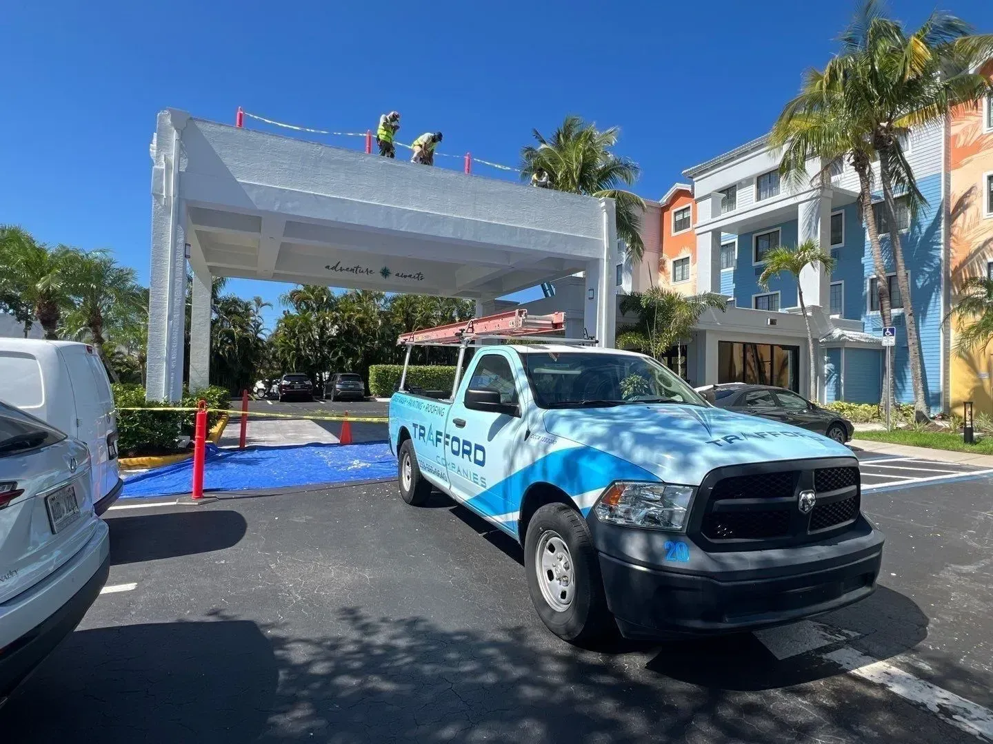 Blue pickup truck under a white event arch on a sunny street, with workers on top and palm trees nearby