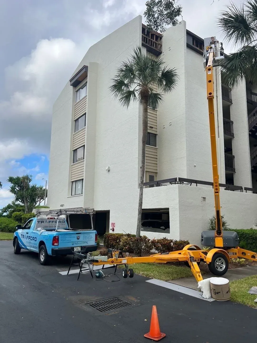 Utility crew trimming palm trees beside a white apartment building, with a blue truck and lift boom nearby.