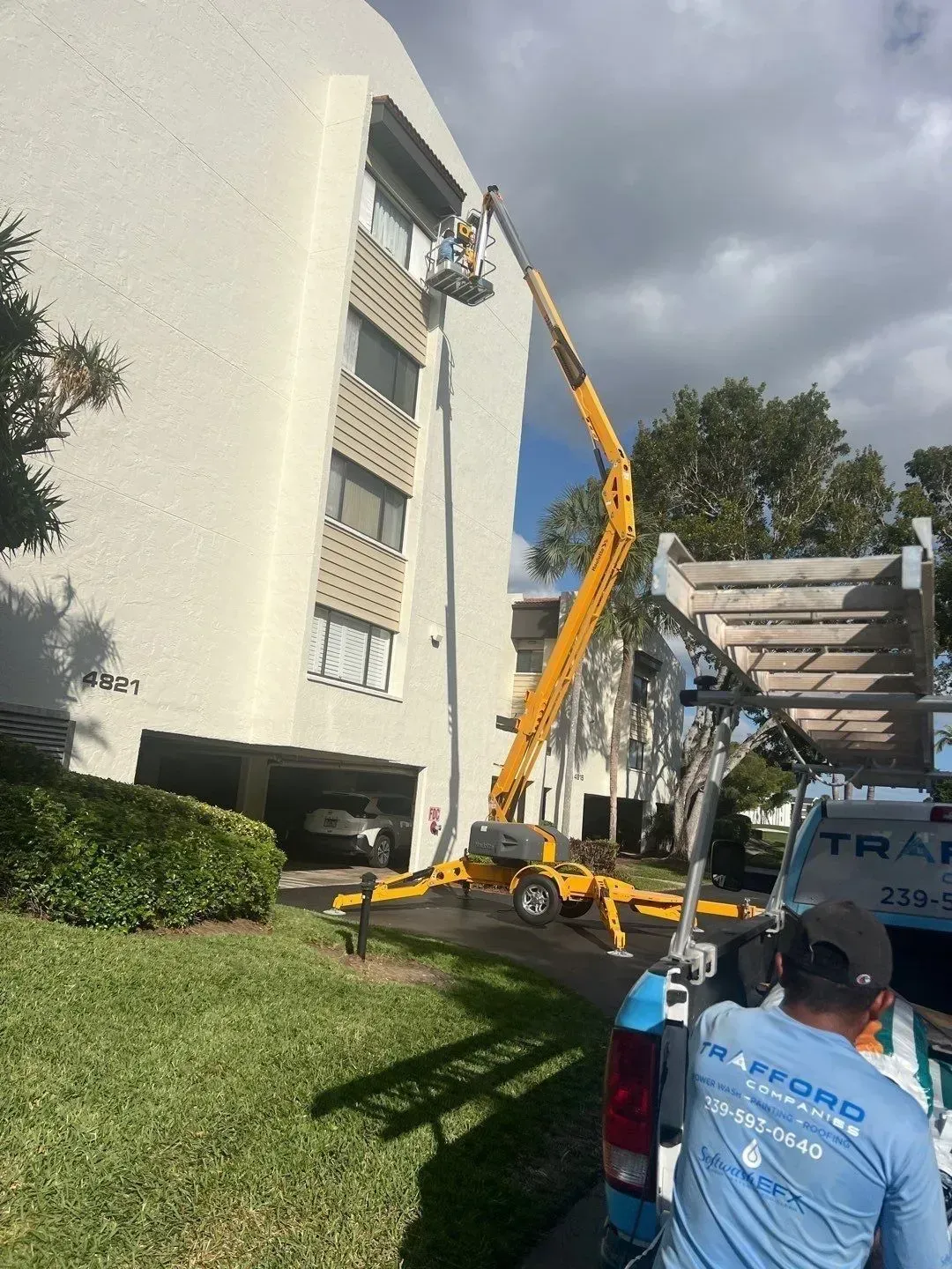 Crane truck lifting workers beside a white apartment building under cloudy skies.