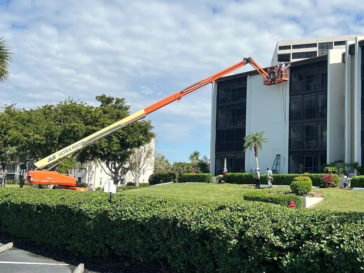 Orange-and-white cherry picker lifting workers beside a white building and hedge on a sunny day
