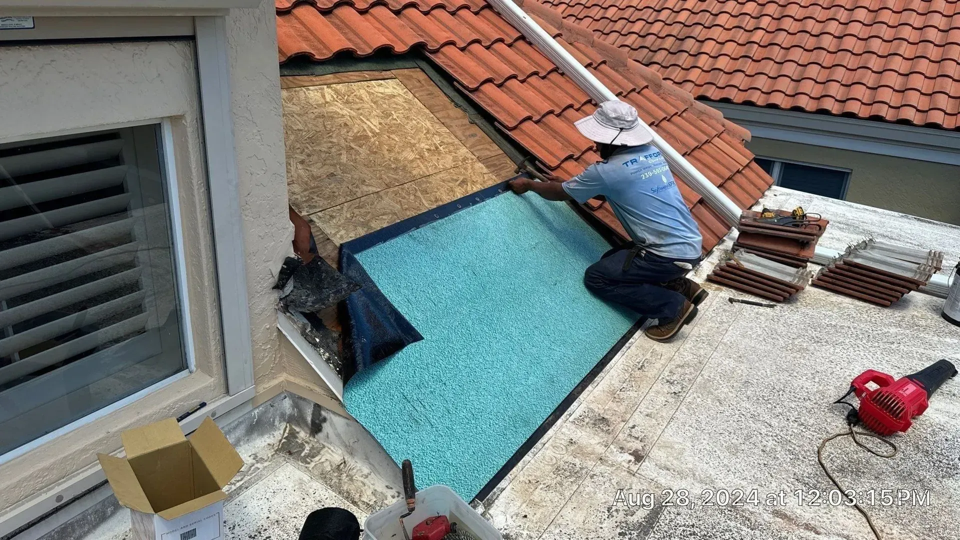 Worker on a rooftop installing blue underlayment beside terracotta tiles and a skylight.