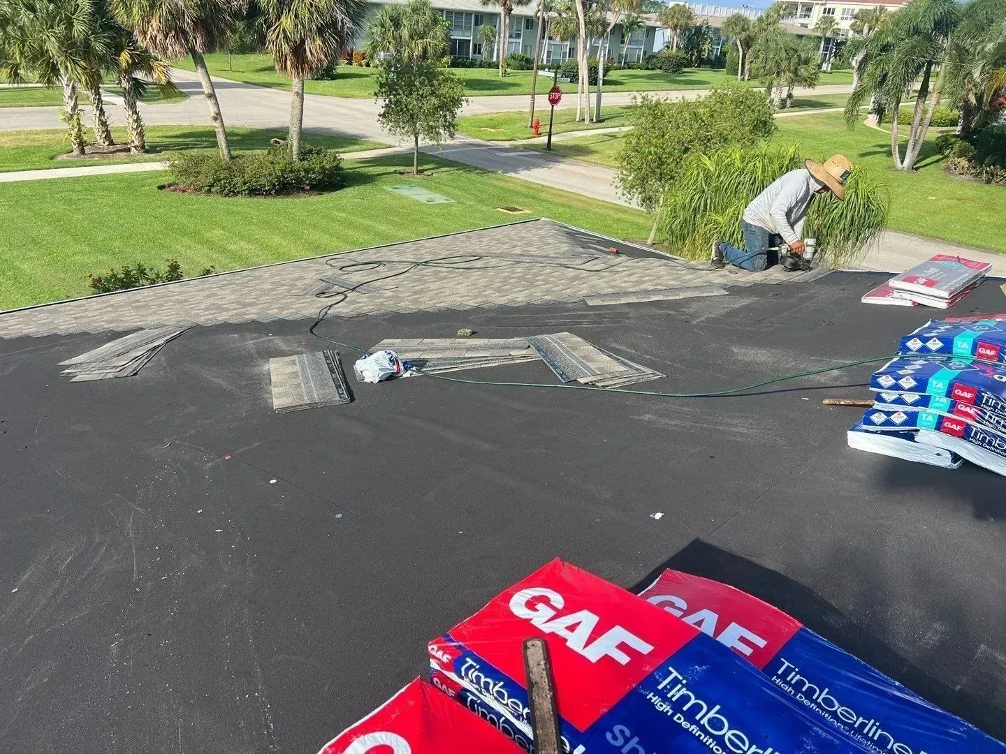 Workers installing dark roofing material on a flat roof beside a grassy park and trees.