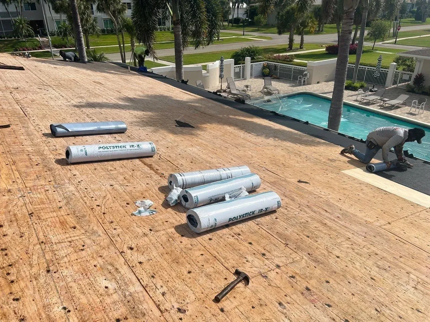 Roofing materials and tools on a sloped roof beside a pool, with workers in the background.