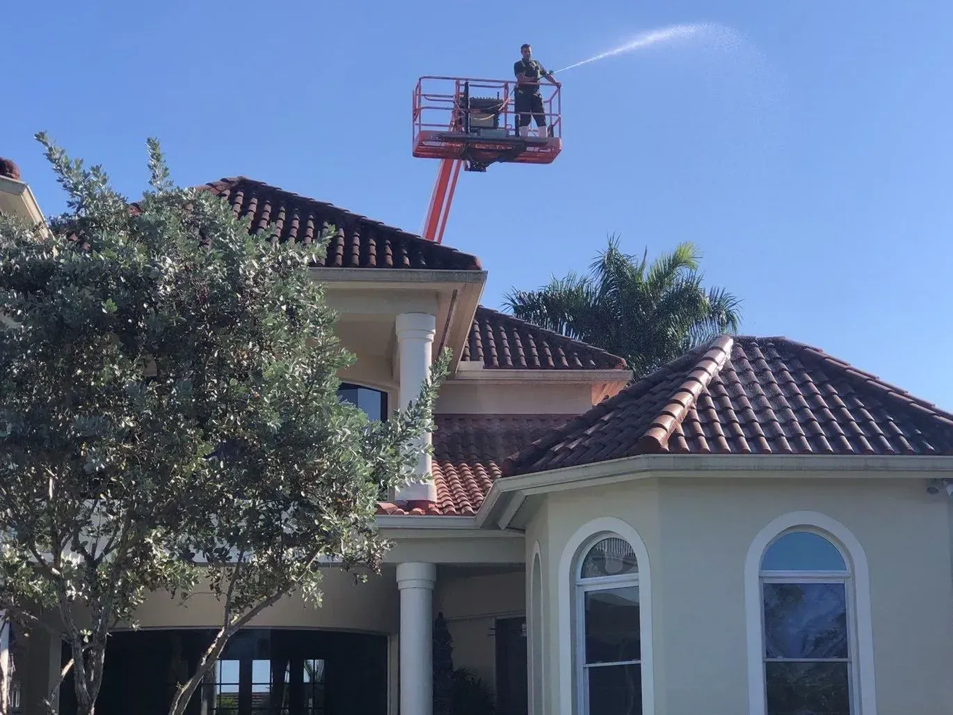 Worker in an orange lift trimming a house roof beside a tree under a clear blue sky