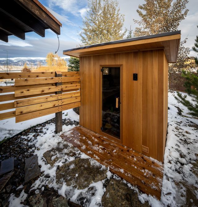 Wooden sauna in snowy outdoor setting, with attached wooden fence and small porch.