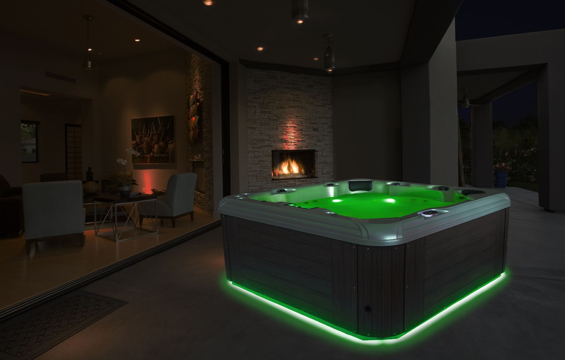 Hot tub illuminated with green lights, on a patio at night, with a fireplace and view of a house interior.